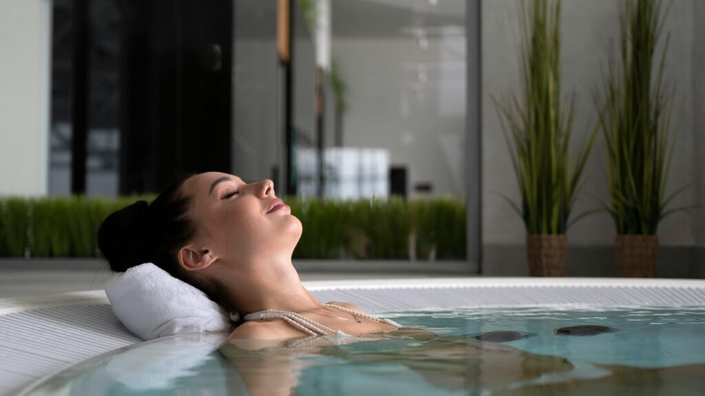 A woman relaxes in a spa pool, eyes closed, with a tranquil expression, surrounded by indoor plants.
